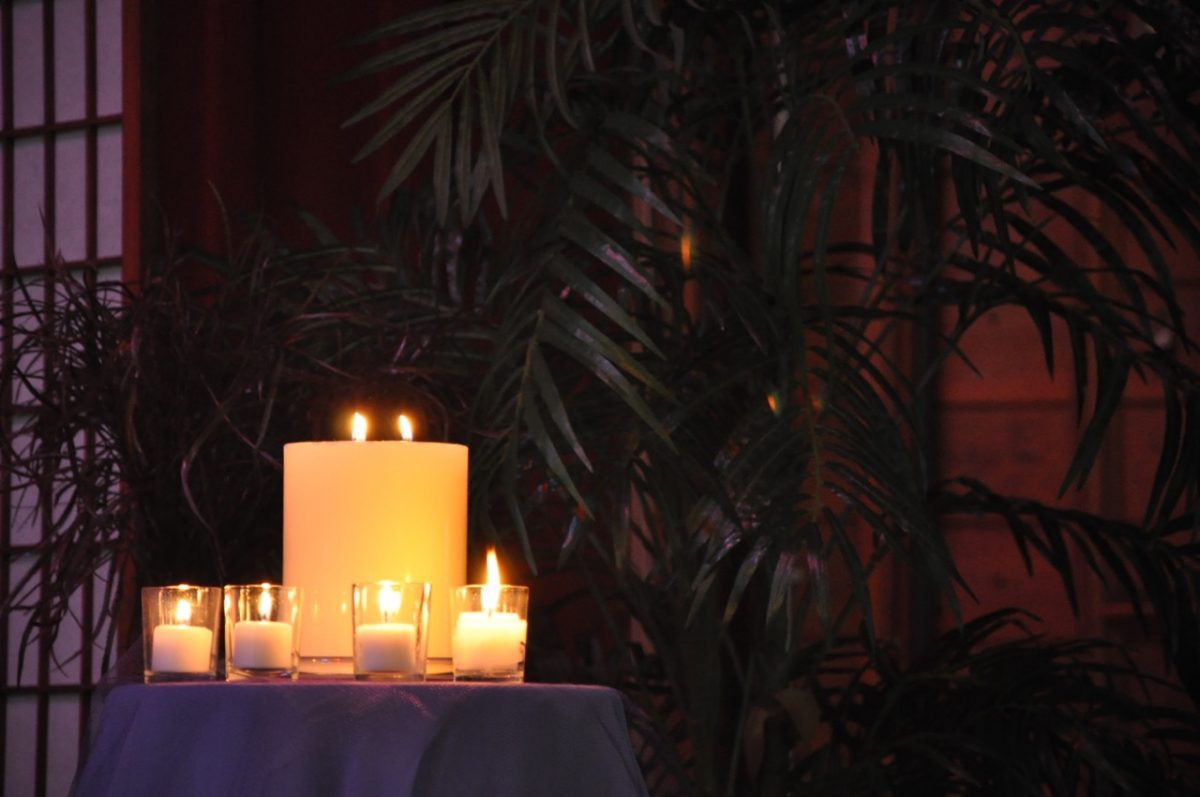 4 Advent votives lit surrounding the Christmas Eve lit pillar candle on a purple table cloth in front of some indoor trees.